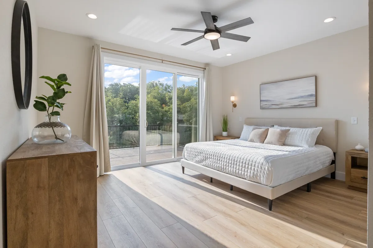 A serene bedroom with a comfortable bed positioned to enjoy direct views through a large sliding glass door, which opens onto a balcony or deck. The room is bathed in natural light, featuring stylish nightstands, a lush plant, and tasteful decor that evoke tranquility and connection to the lake just beyond.