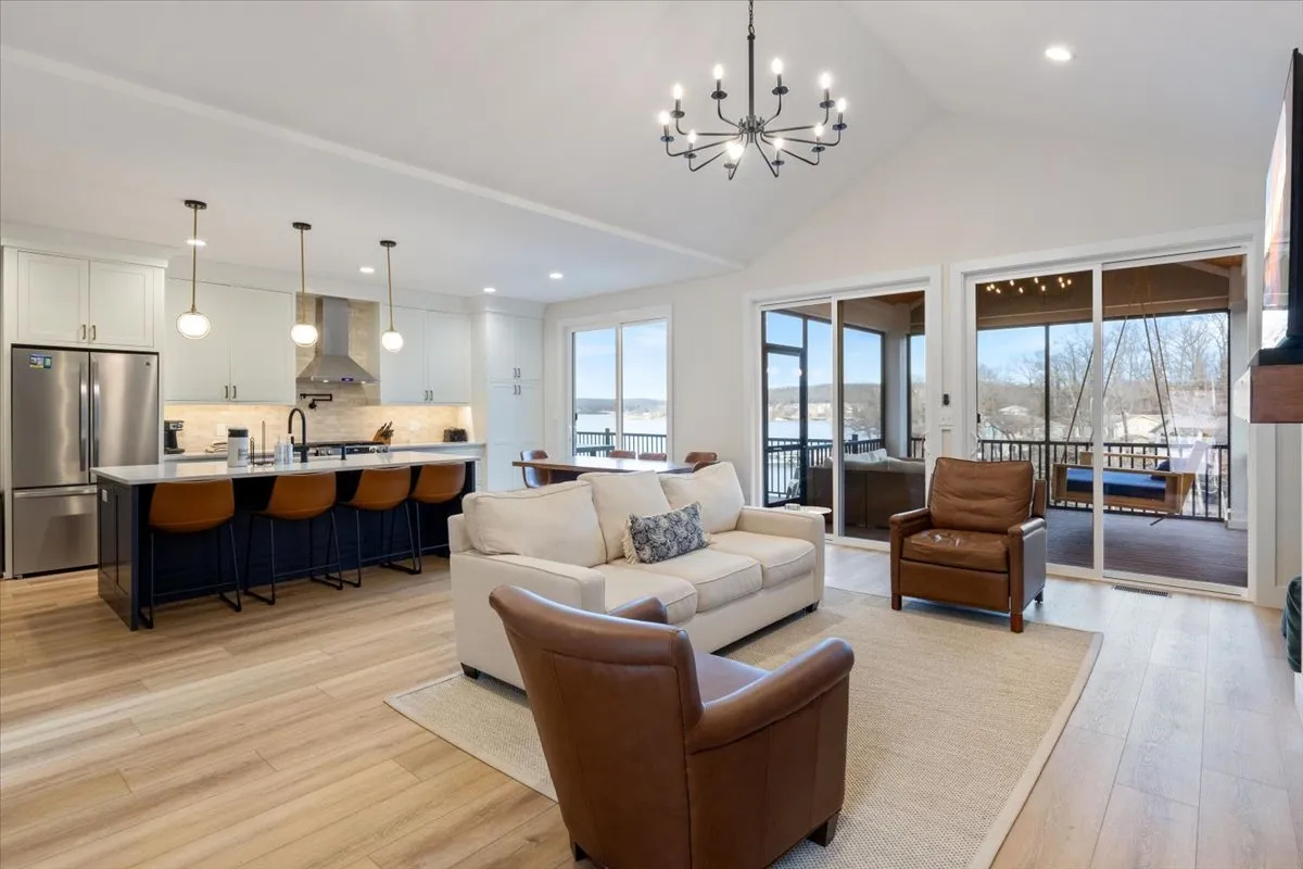 A wide-angle view of an open concept living area featuring a stylish kitchen island with bar seating, a plush sofa and armchairs, and expansive windows that flood the space with natural light. A striking chandelier completes the sophisticated, welcoming look.