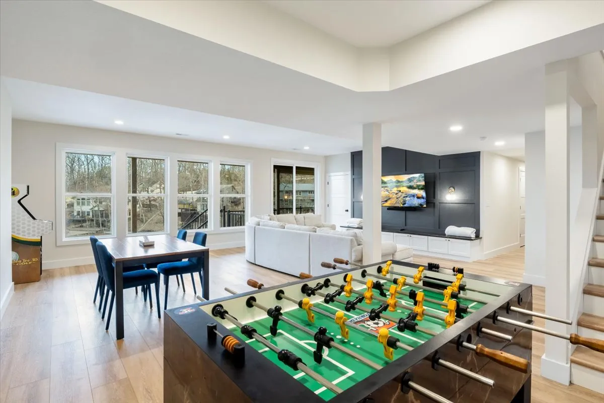 This image captures a vibrant lower-level entertainment area featuring a foosball table as the focal point, a large TV, spacious comfortable seating area, and big windows that flood the space with natural light. The playful setup and relaxed decor highlight opportunities for group fun and relaxation.