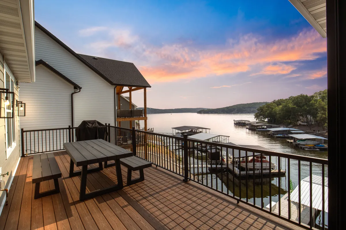 The image showcases the exterior of a modern lakefront home at sunrise or sunset, with sweeping views of the tranquil lake, a spacious outdoor dining area set for al fresco meals, and boathouses along the shoreline. The scene evokes easy access to the water, relaxation, and gathering with loved ones.