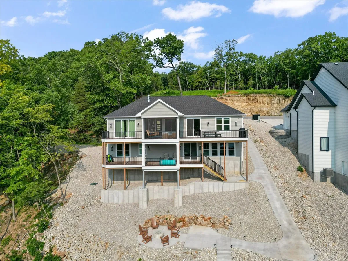 An exterior shot highlighting the two-story house's walkout lower level, a cozy fire pit area arranged with seating, and the scenic lake backdrop. The setup invites guests to step outside for evening gatherings around the fire and lakeside relaxation.