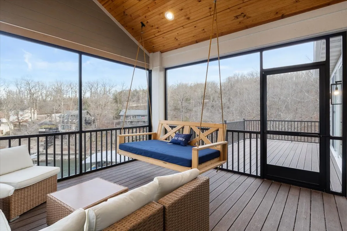 A tranquil screened-in upper deck featuring a cozy swing bed, comfortable wicker seating, and a view through large windows. The wood-paneled ceiling and inviting decor make it a perfect spot for relaxed mornings or spirited evening gatherings.