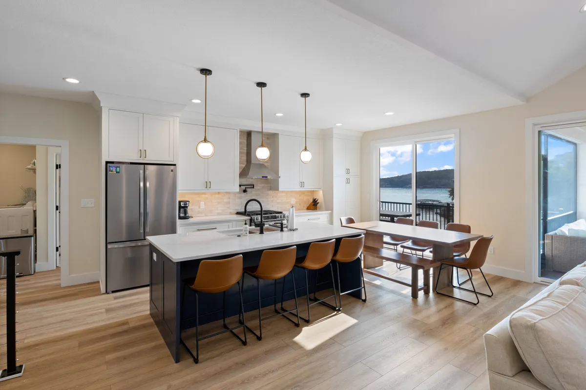 A bright kitchen and dining area with a large island, elegant dining table, and panoramic windows revealing stunning lake views. The space is airy, modern, and ideal for family-style meals or entertaining guests.