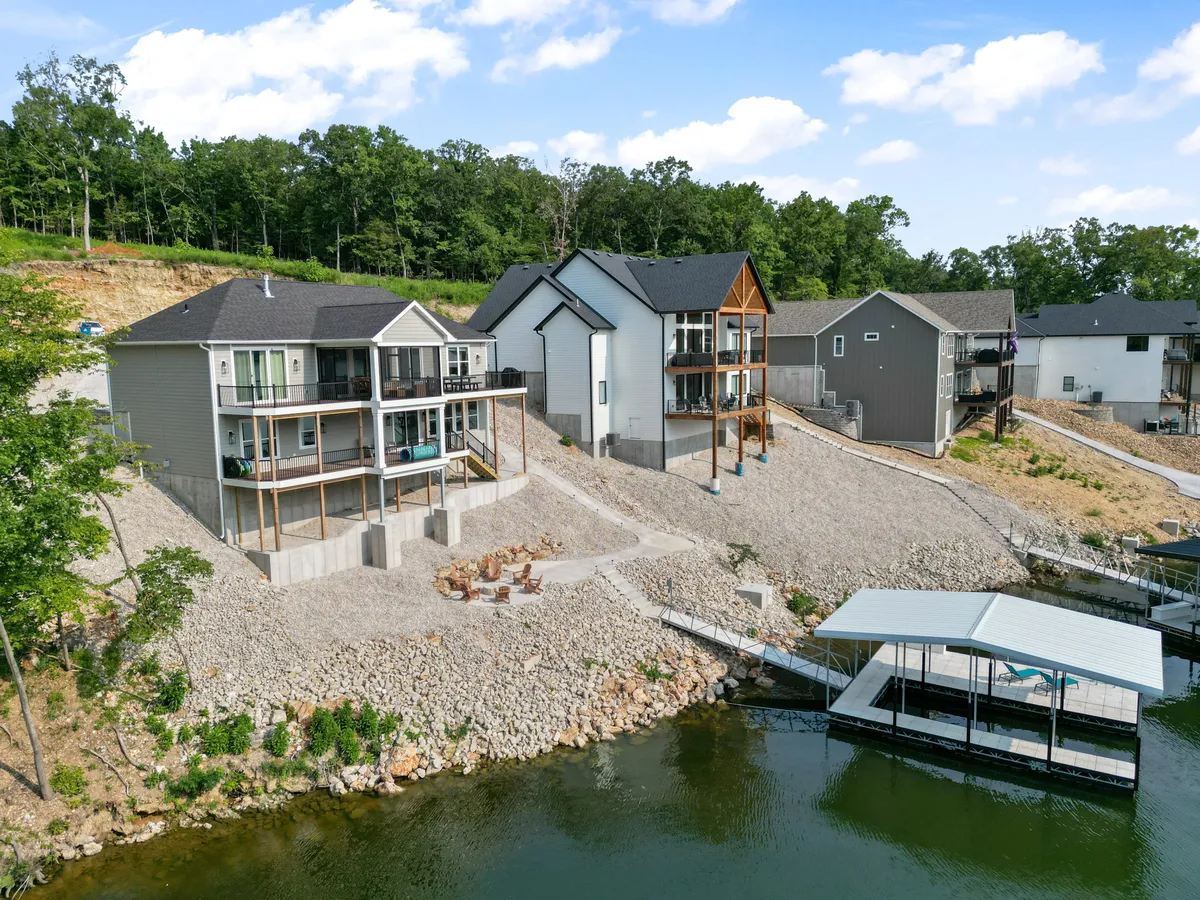 A stunning lakeside scene featuring modern homes with sleek architectural lines, an outdoor seating area, and a boat dock stretching into the calm water. Lush greenery surrounds the lakefront, creating a sense of tranquility and exclusivity.