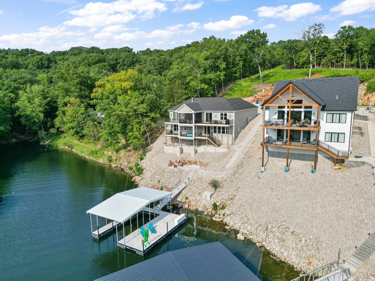A panoramic view of modern lakefront homes framed by leafy trees, with a boat dock extending over a glassy water surface. The surroundings exude peace and the possibility for outdoor recreation, from boating to evening bonfires.